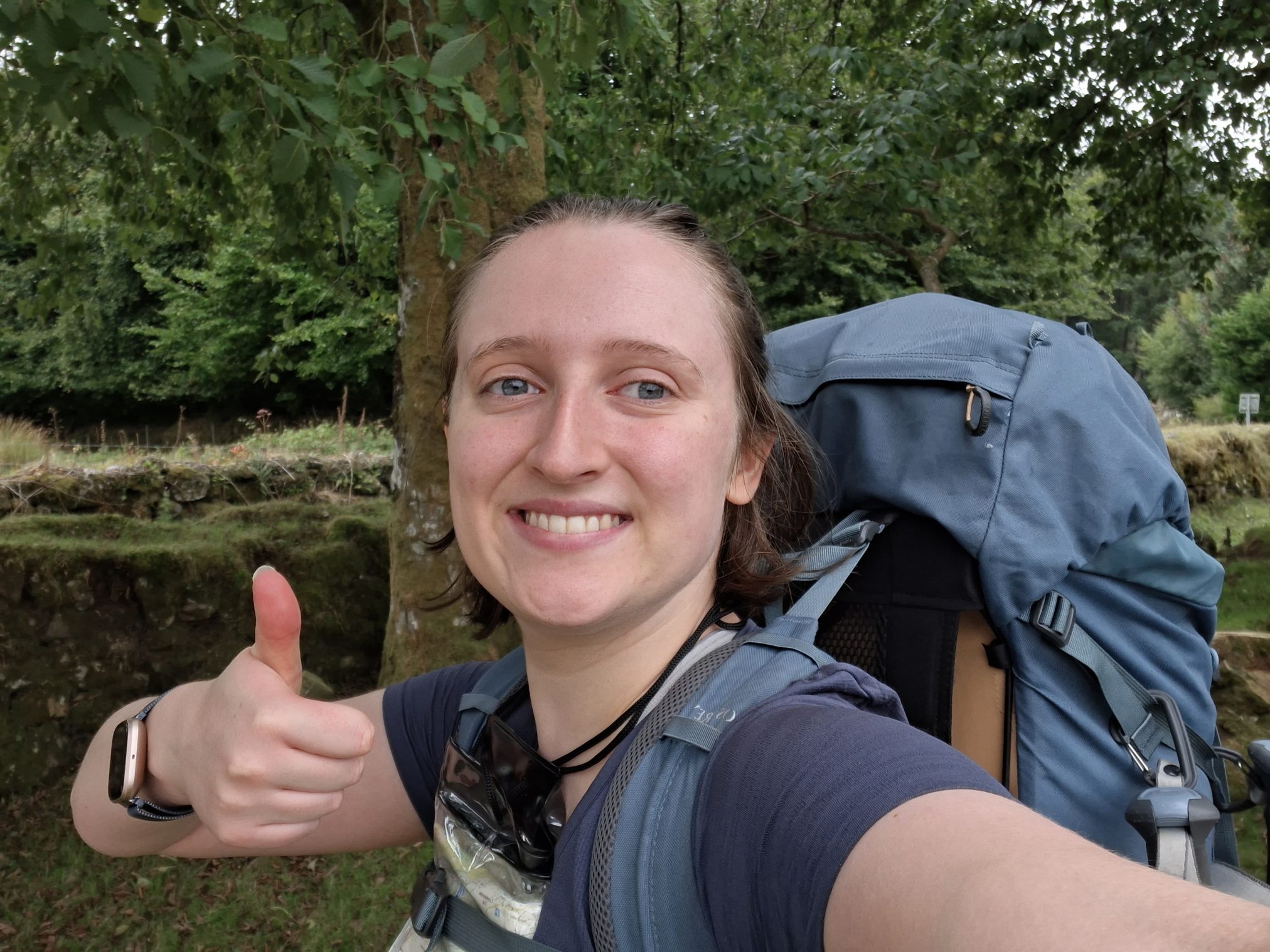 Selfie of Anna, smiling and giving a thumbs up, wearing their backpack in nature.